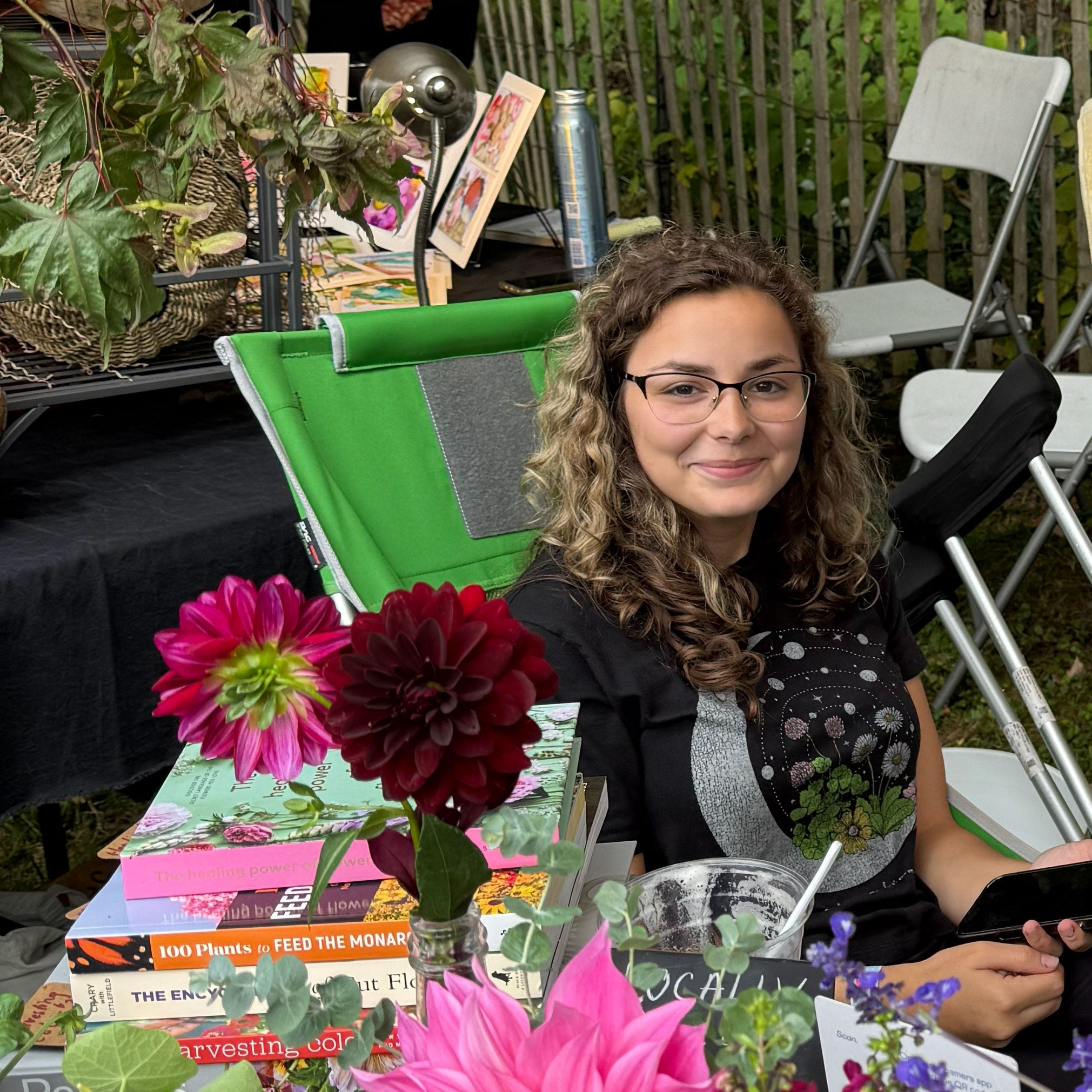 Woman with glasses sitting at a table with books and flowers, smiling outdoors.