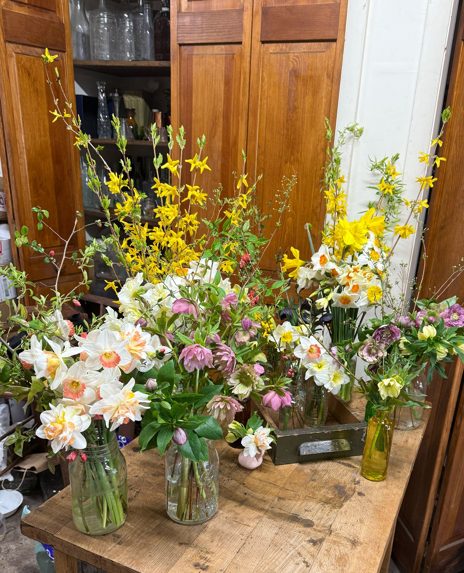 Bouquets of flowers in glass vases on a wooden table with a wooden cabinet in the background.