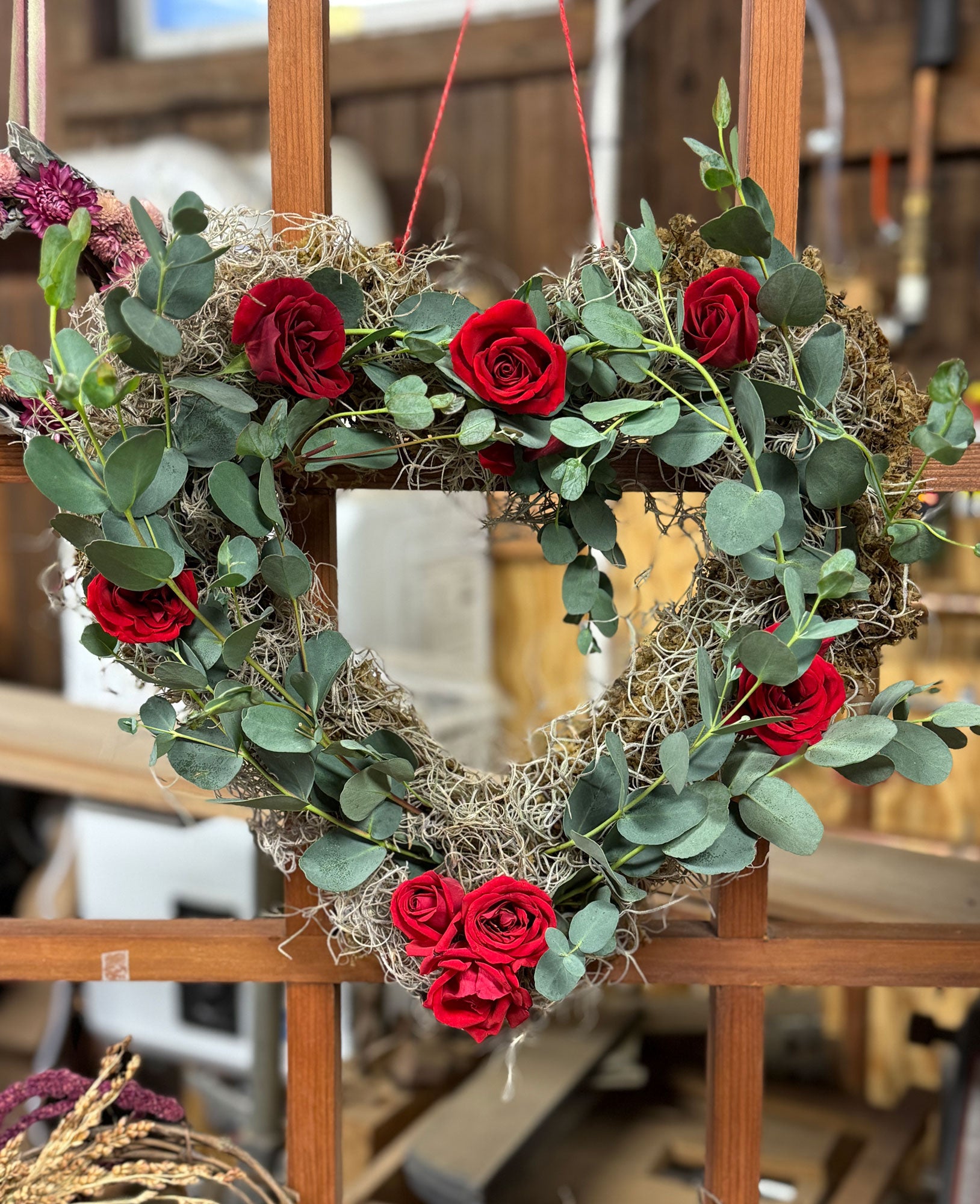 Heart-shaped wreath with red roses and greenery on a wooden stand.