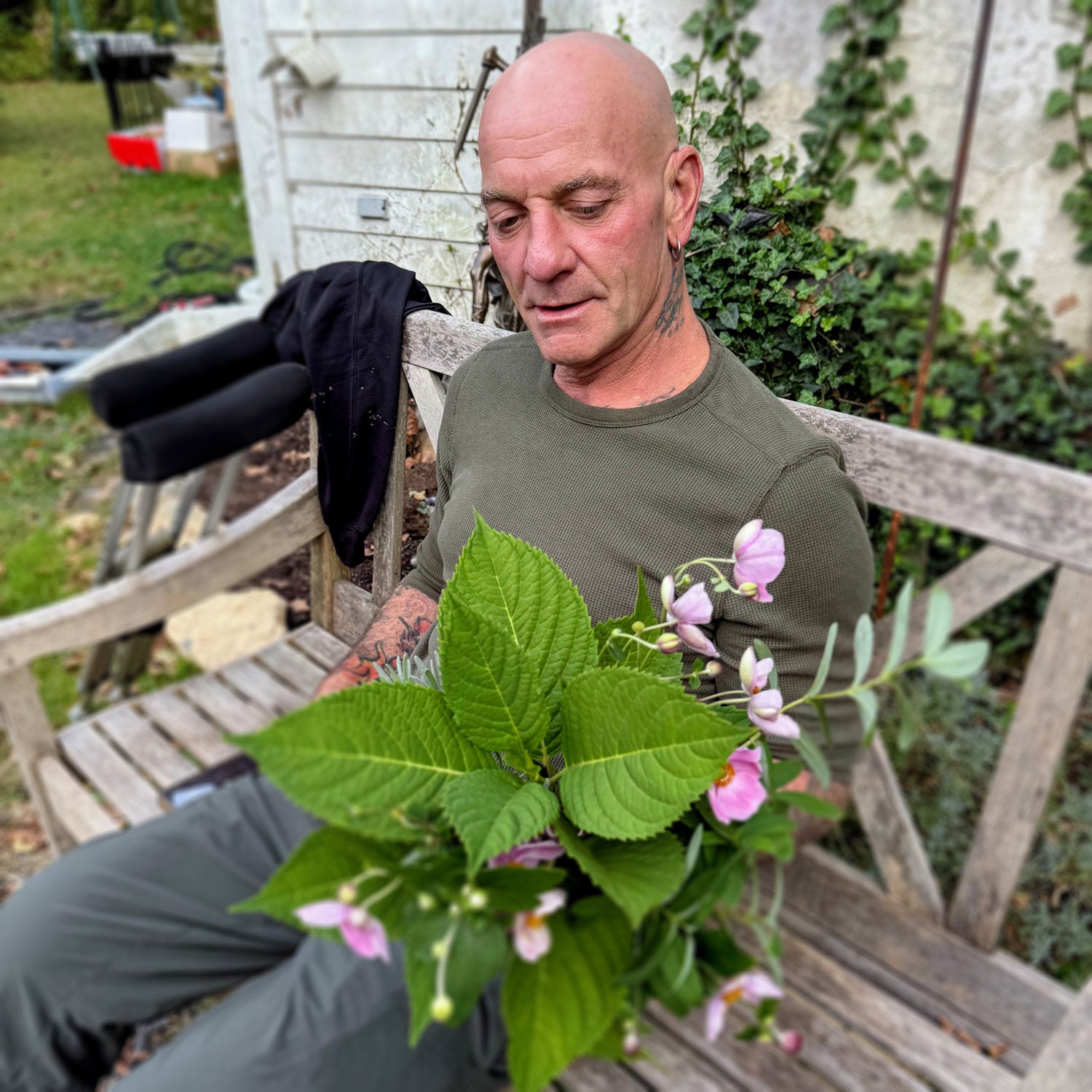 Man holding a plant with pink flowers and green leaves, sitting on a wooden bench outdoors.